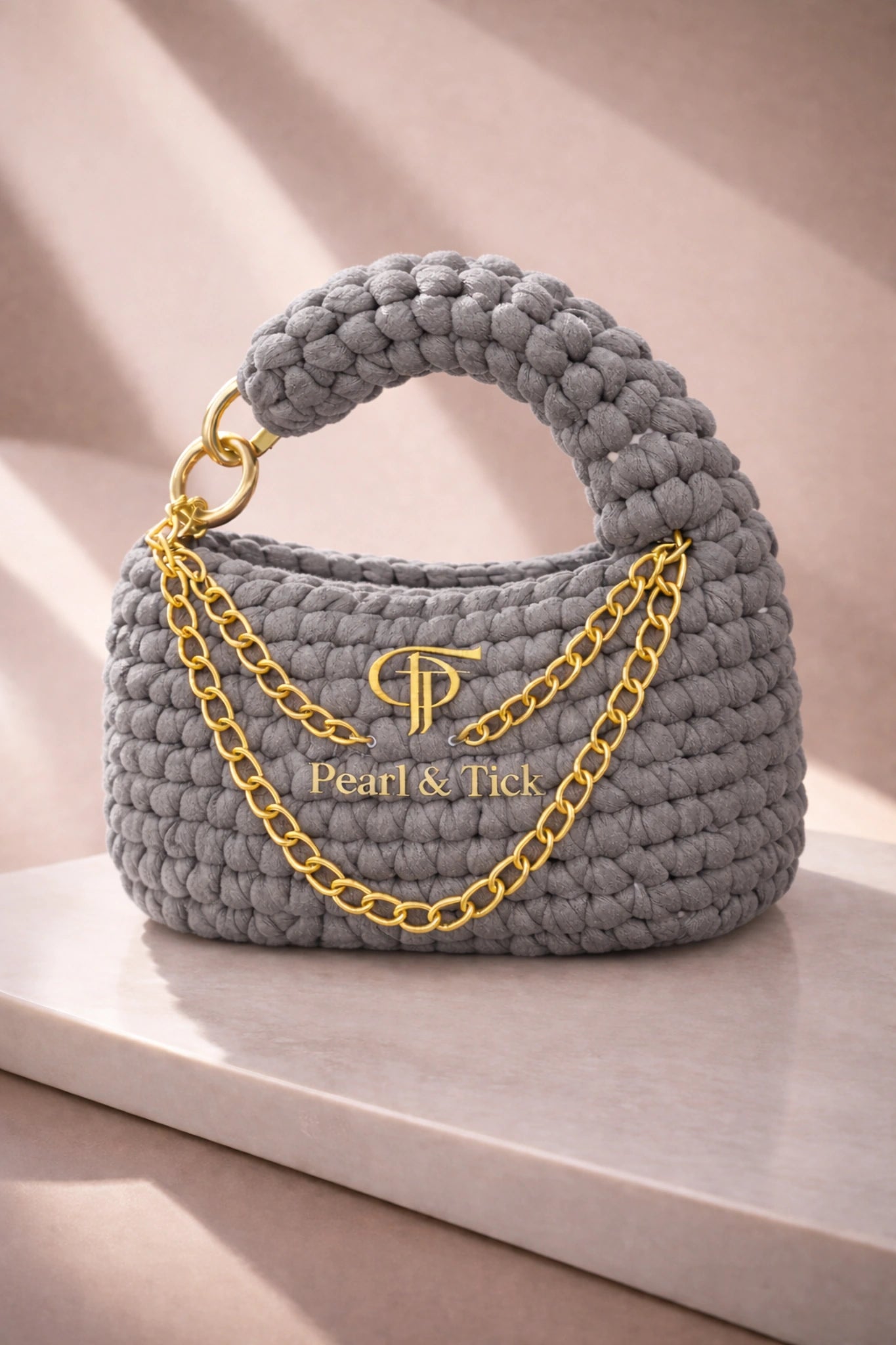 Borsa grigia con catena dorata e marchio 'Pearl & Tick' su una superficie chiara.