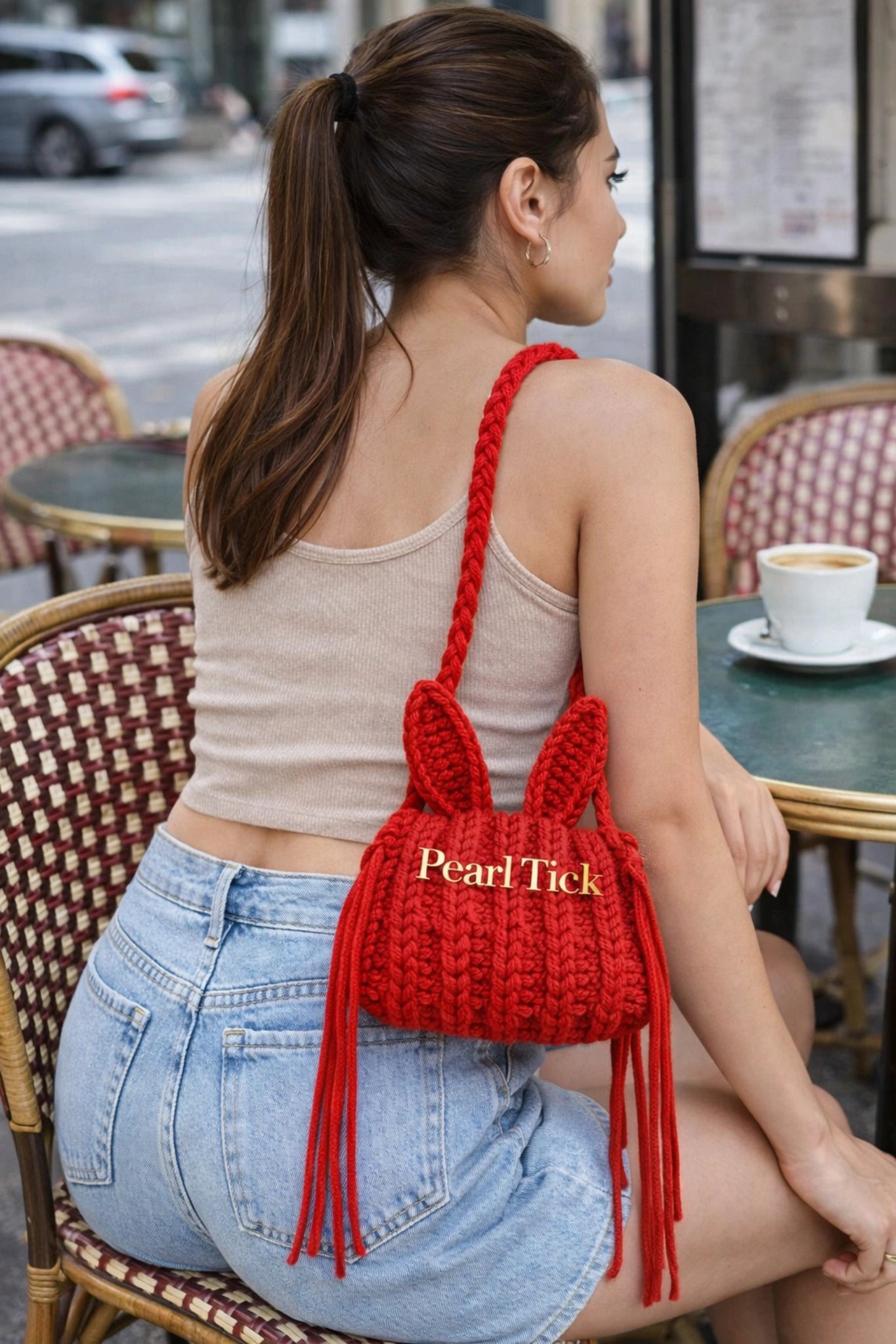 Woman sitting at an outdoor cafe table with a red knitted bag on her shoulder.