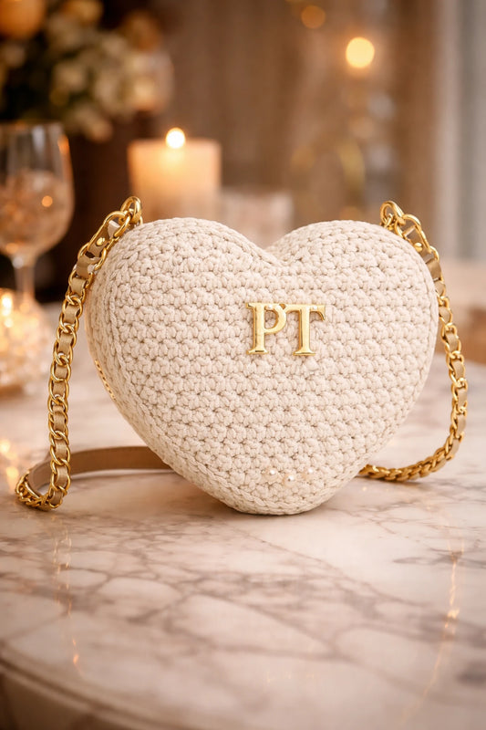 Heart-shaped white handbag with gold chain and 'PT' initials on a marble surface.