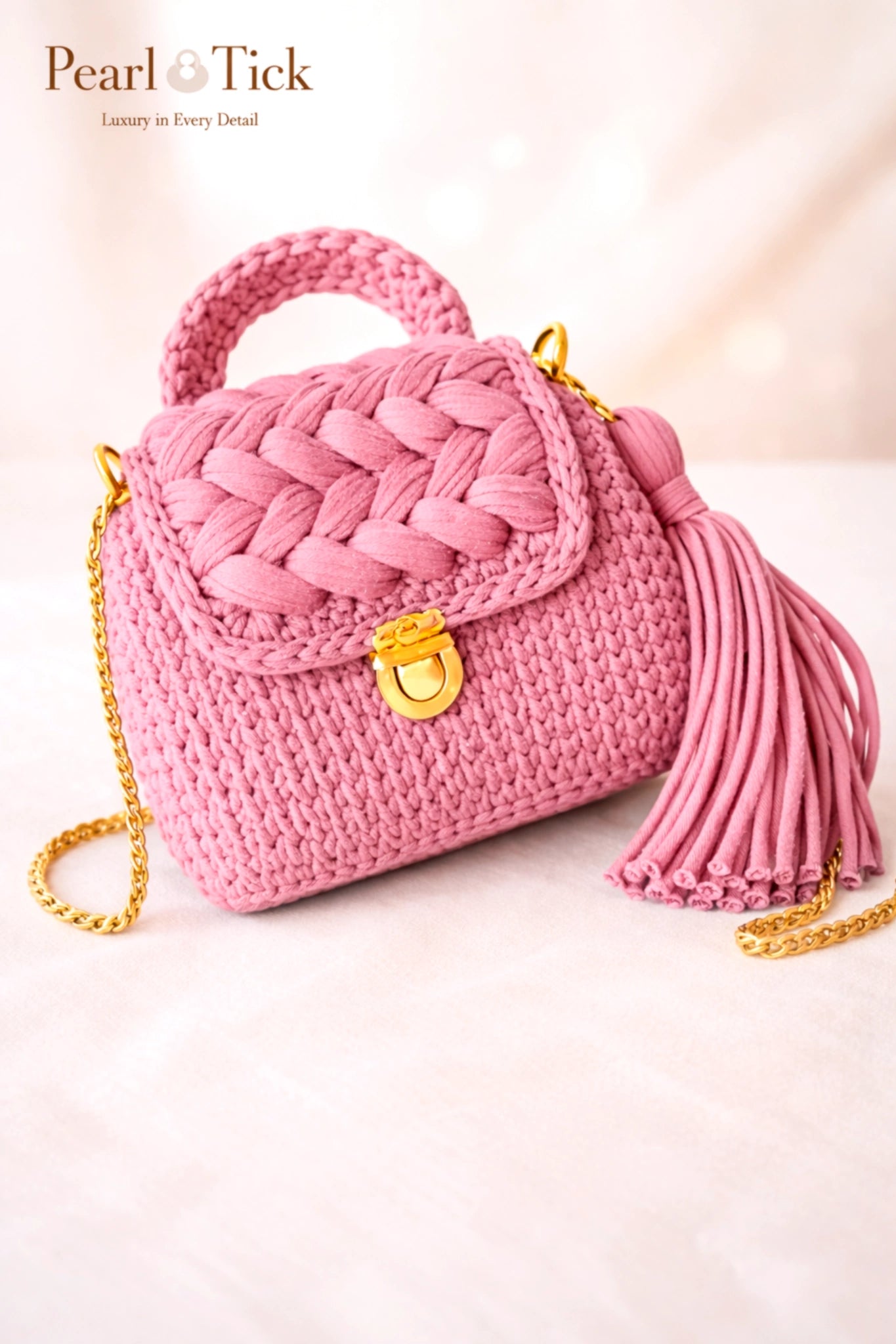 Borsa a mano rosa intrecciata con nappa e catena dorata su sfondo rosa chiaro, con il marchio «Pearl & Tick».