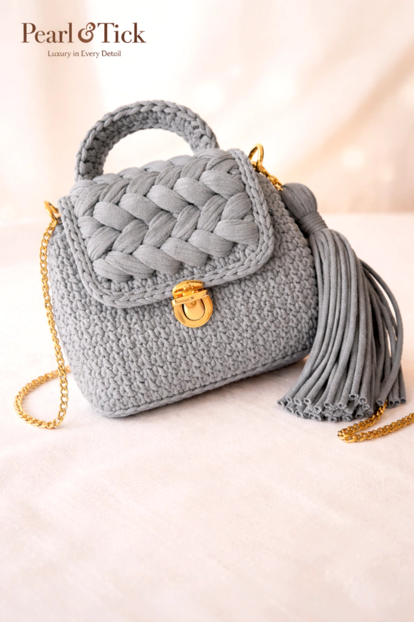 Borsa a mano grigia in tessuto con catena dorata e nappina su sfondo chiaro, con il marchio «Pearl & Tick».