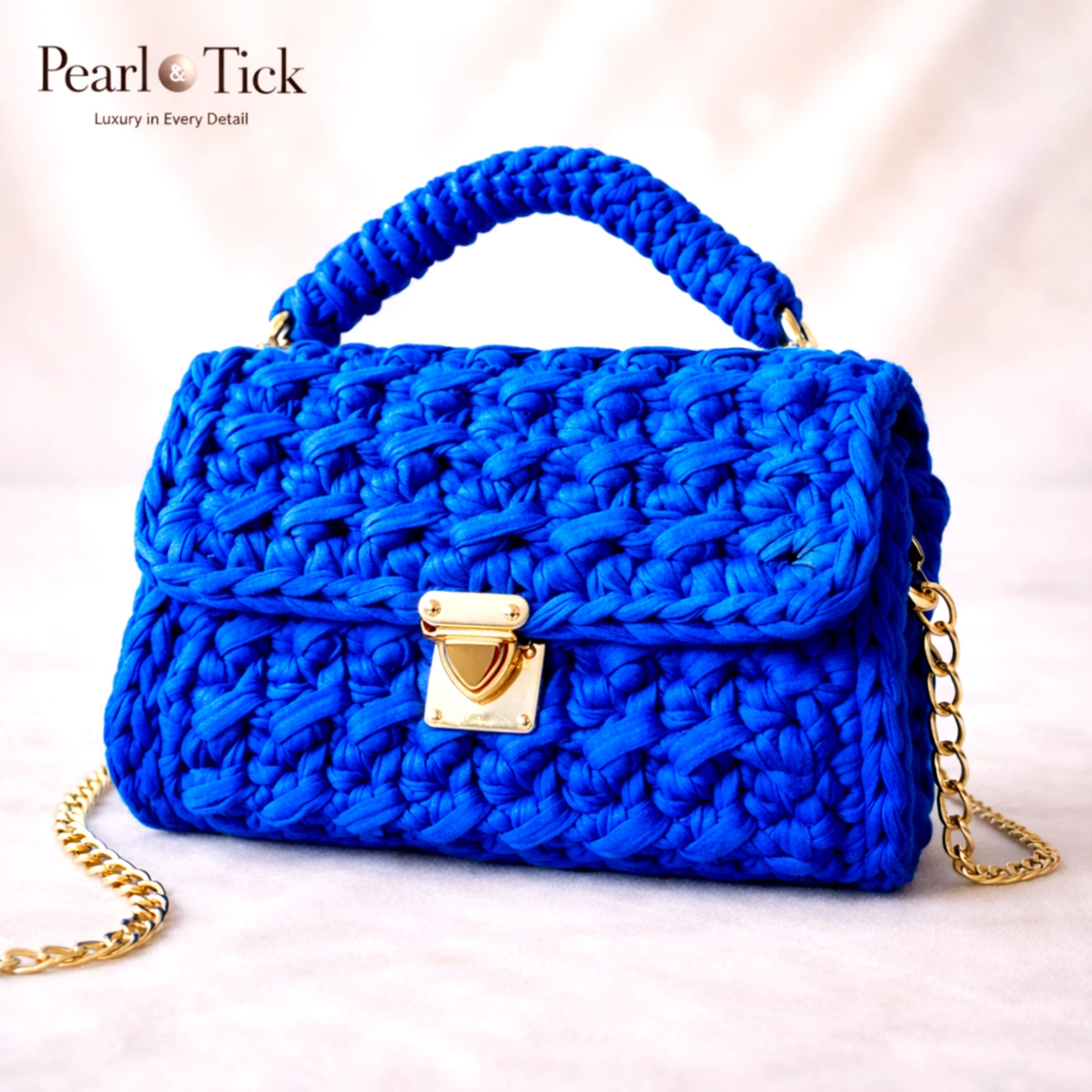Sac à main tressé bleu avec chaîne et fermoir dorés sur un fond rose clair, estampillé 'Pearl Tick'.